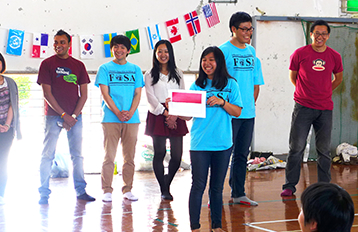 International students lined up in a hall with flags from various countries at the back. A lady stands in front giving a speech.