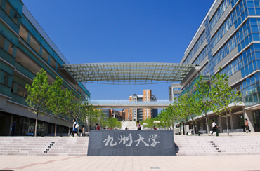 九州大学伊都キャンパス正門。九州大学と彫られた銘板の背後に九州大学の施設が広がる風景